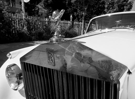 Magazine style fashion photoshoot in Los Angeles. Portrait of the Bride and Groom in a kiss reflected in a Rolls Royce classic car limousine. Brides veil is blowing in the wind.